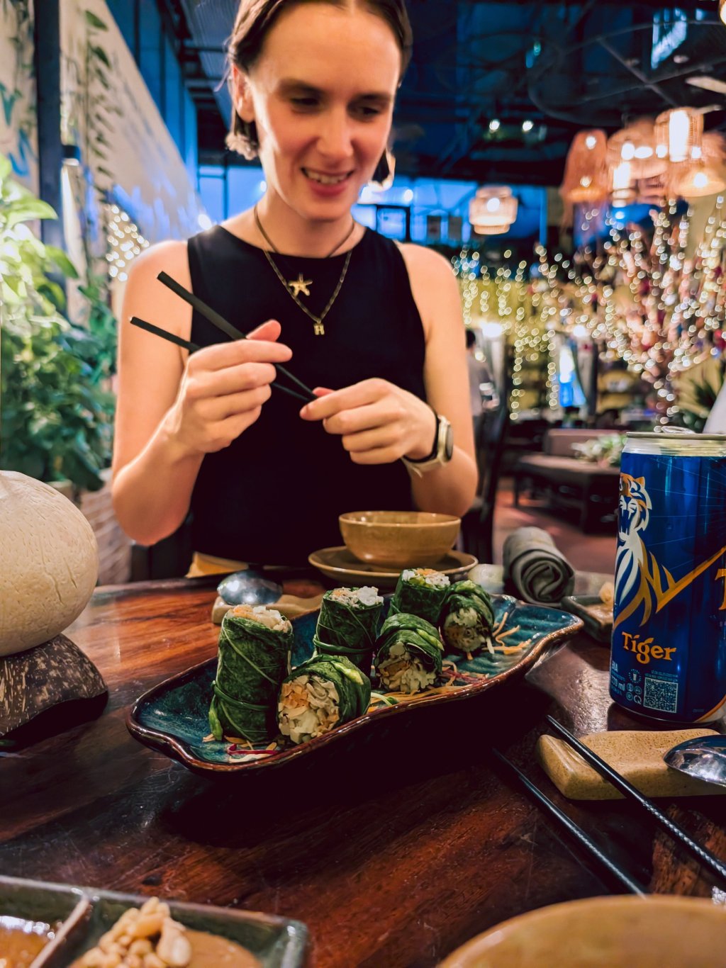 A person is preparing fresh spring rolls at a wooden table in a bustling Ho Chi Minh City setting.