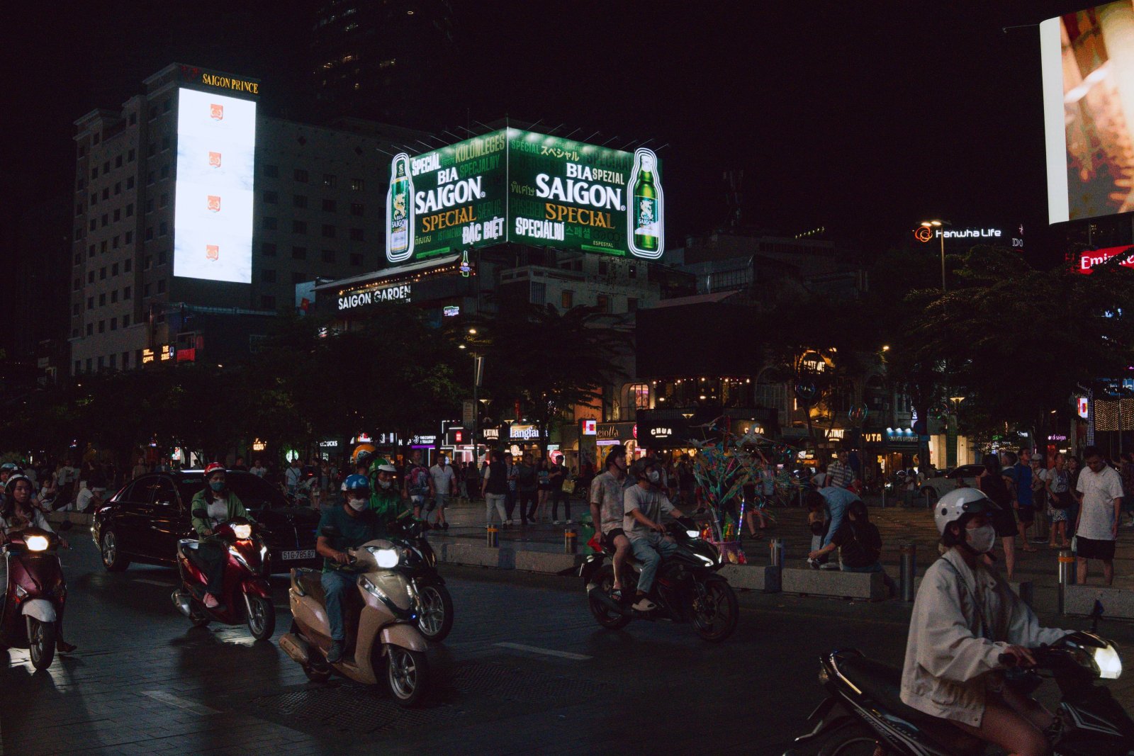 Night street scene in Ho Chi Minh City featuring illuminated billboards and motorbike traffic.