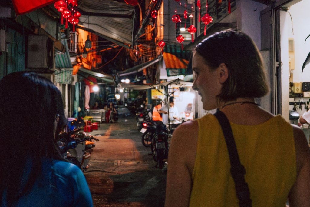 People walking along a narrow city street with scooters and red lanterns at night.