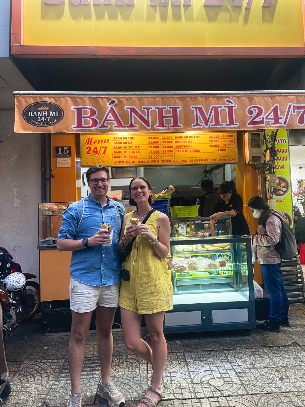 Two people standing in front of a street food stall displaying various items.