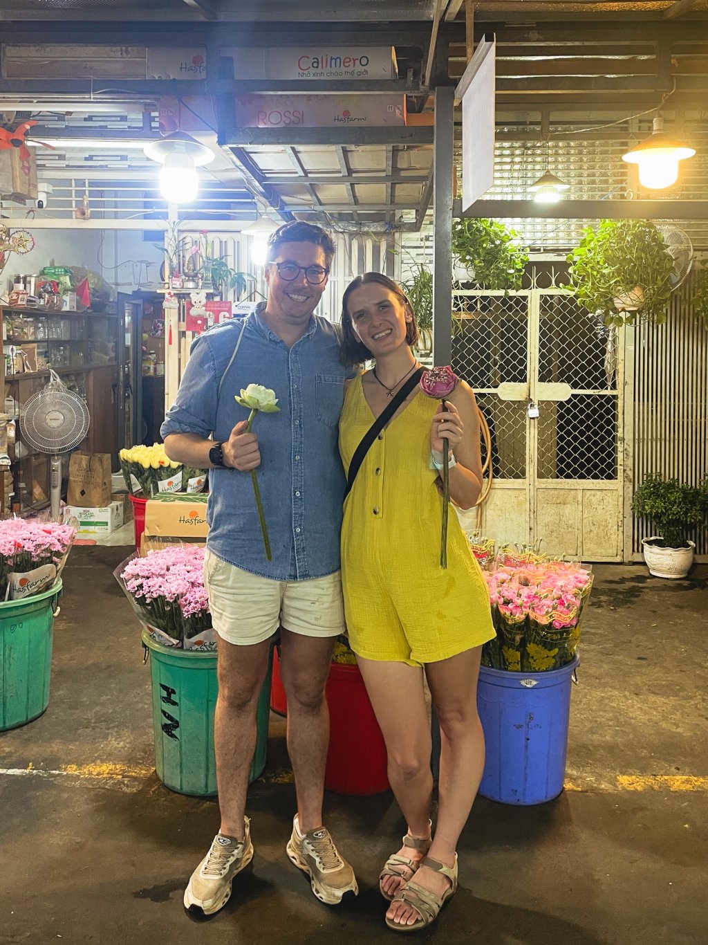 Two people standing in a market setting with flowers and produce.