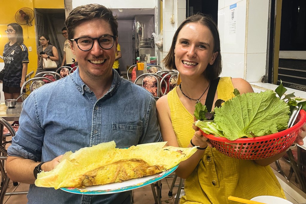 Two people smiling while eating street food in a market setting.