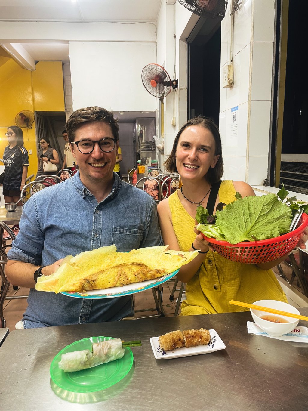 Two people smiling while eating street food in a market setting.