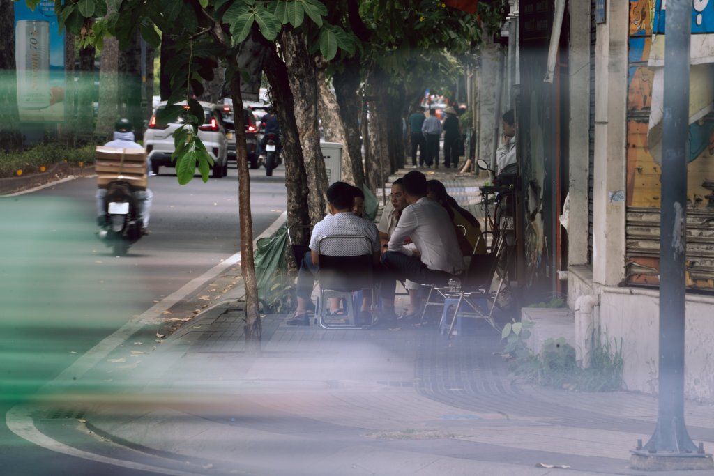 People sitting along a roadside with trees and buildings in a city setting.