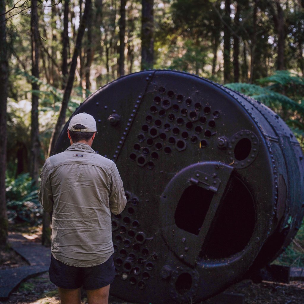 Chris standing before a large, rusted iron boiler resting in a lush, green forest setting.