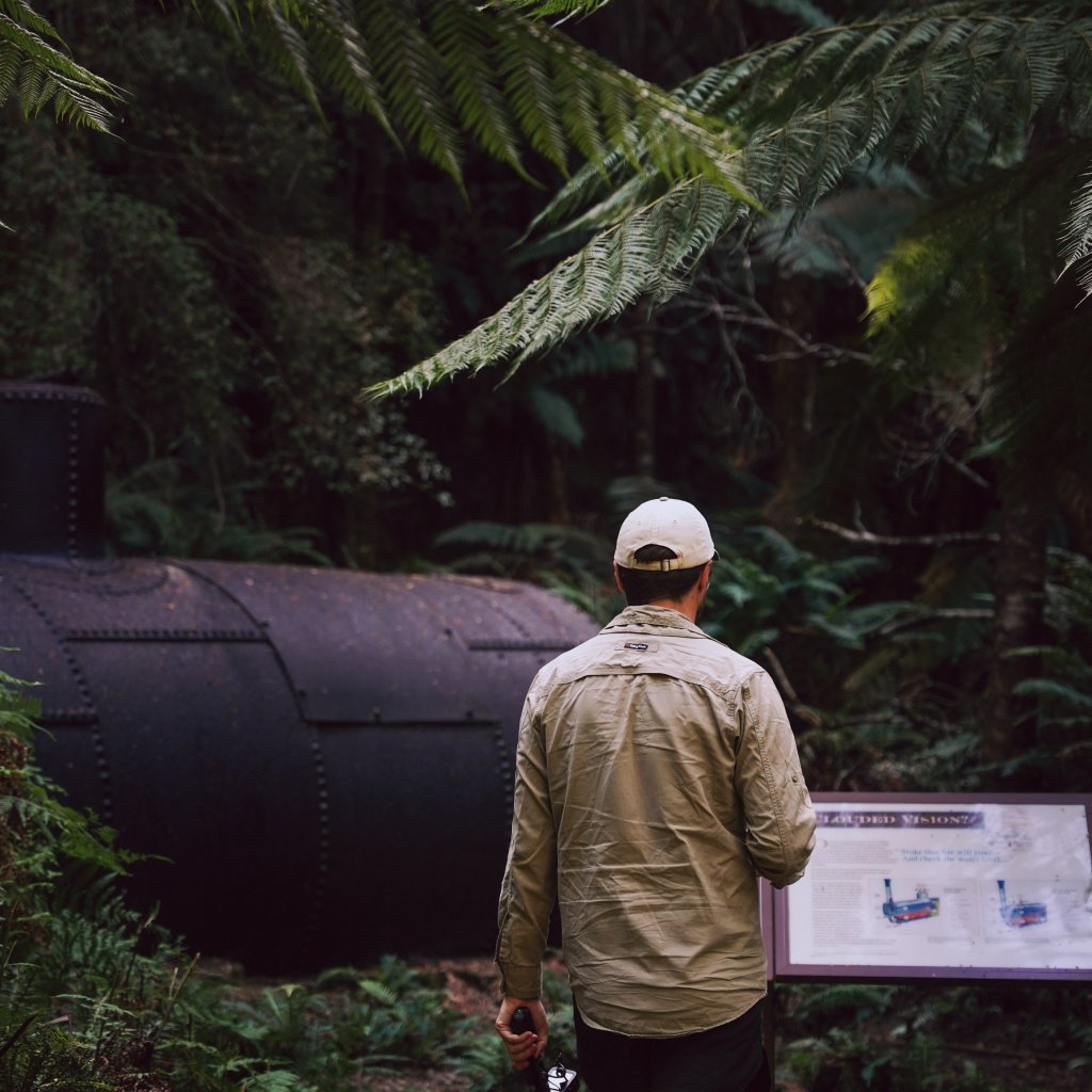 Chris walking past a rusted steam boiler and information sign deep within a lush fern forest.