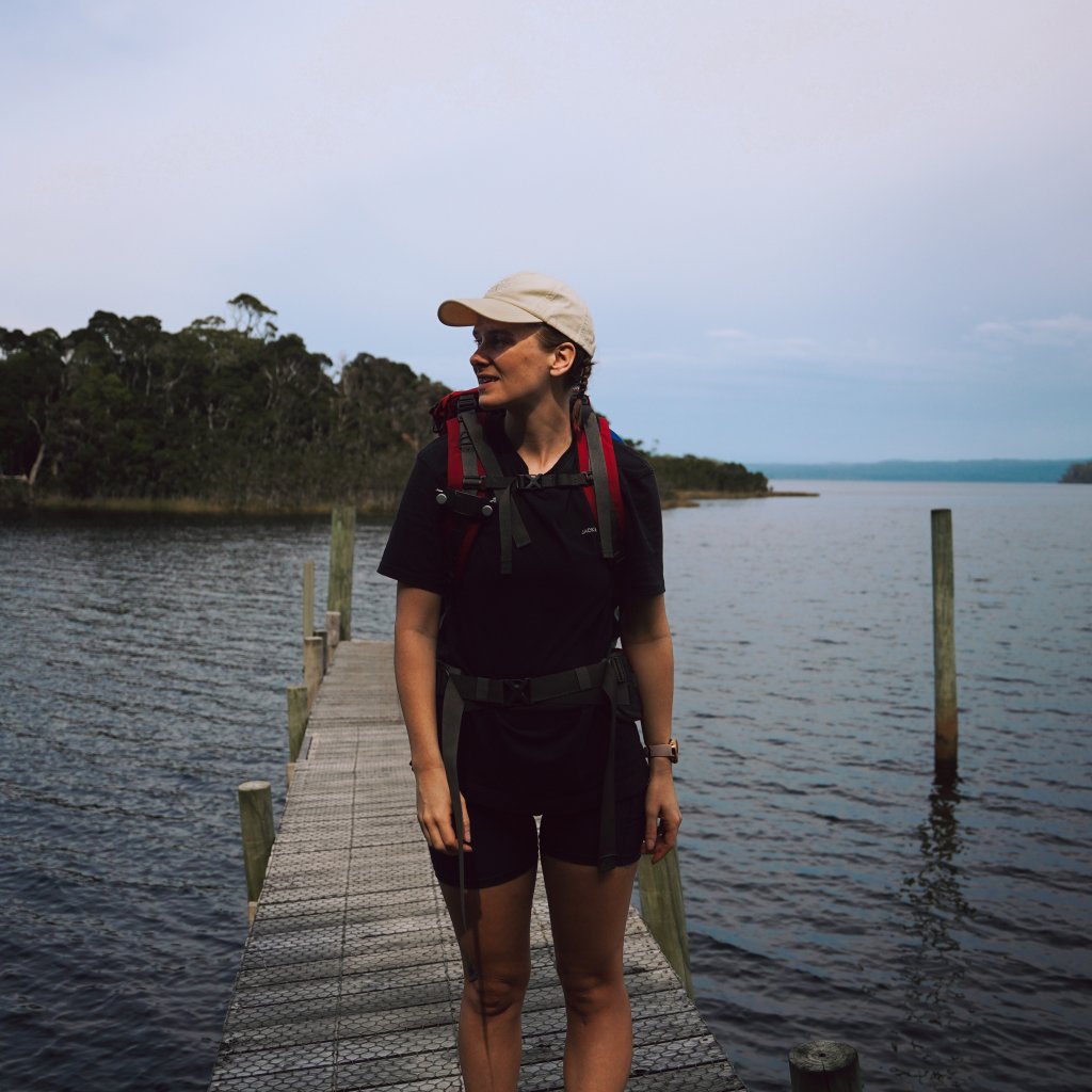 Hannah standing on a wooden jetty overlooking the calm waters and forested shoreline of a quiet bay.