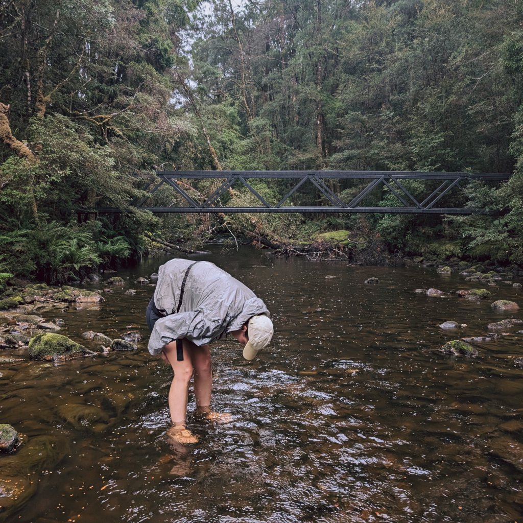Hannah wading through a shallow, rocky river beneath a steel bridge in a lush green forest.