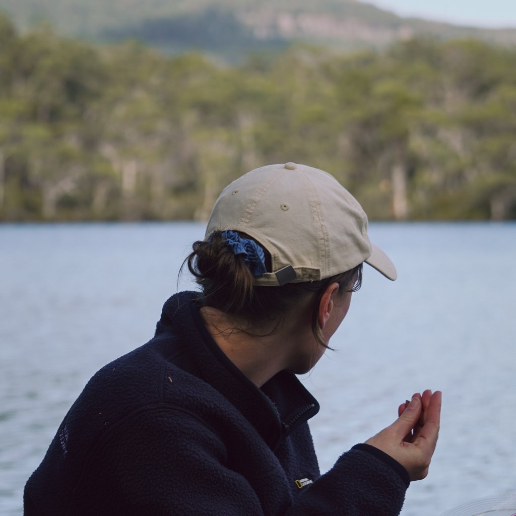 Hannah sitting by the calm water of a lake, wearing a fleece and cap while enjoying a snack.