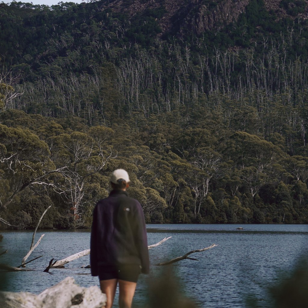 Hannah standing by the calm waters of Lake St Clair, looking toward the rugged peaks and dense forest.