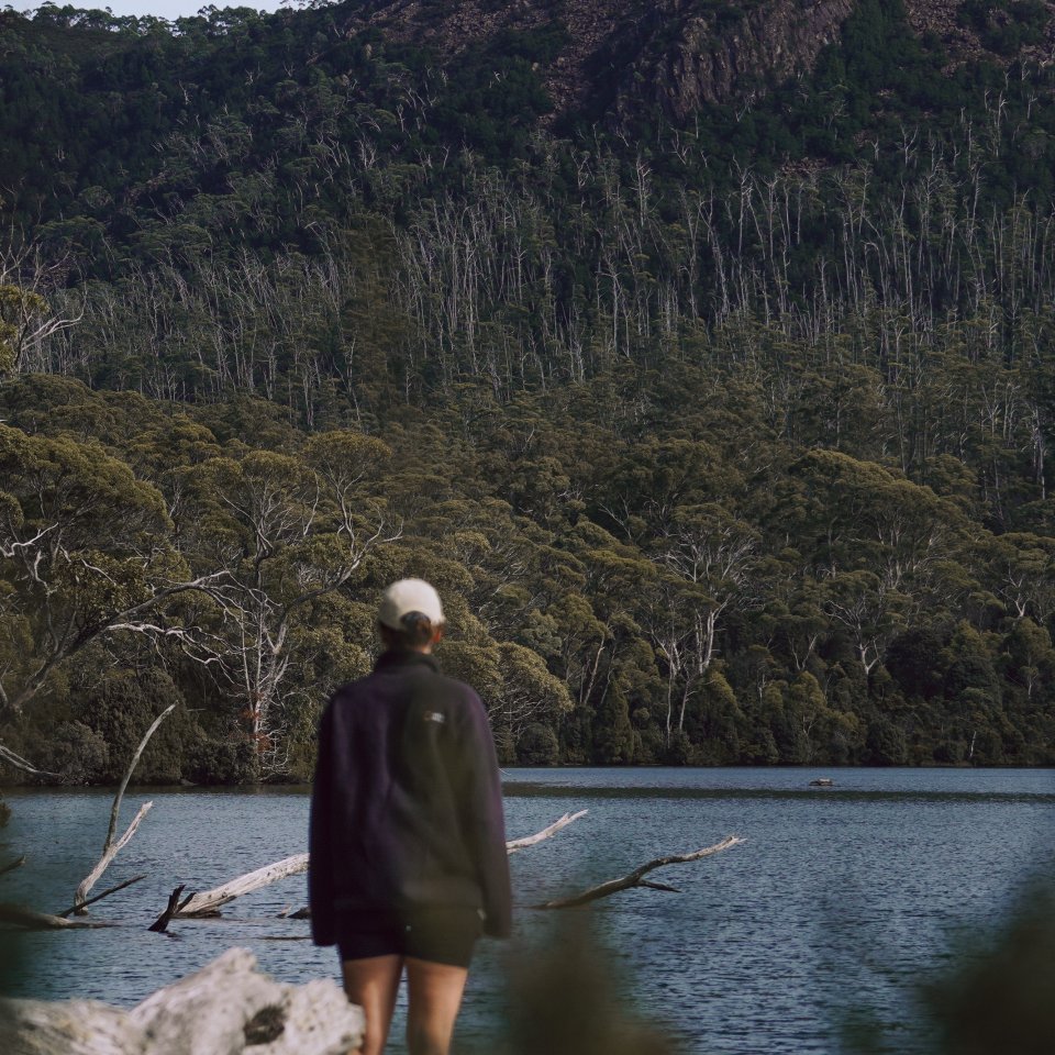 Hannah standing by the calm waters of Lake St Clair, looking toward the rugged peaks of Mount Olympus.