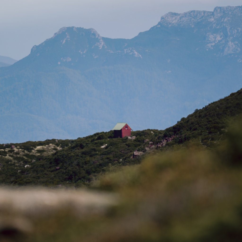 A small red hut nestled on a green hillside with blue mountain peaks in the distance.