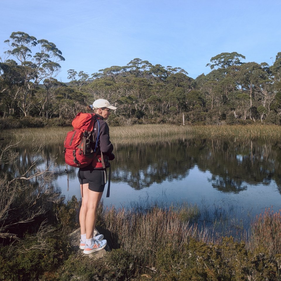 Hannah standing with a red backpack by a still lake reflecting the surrounding eucalyptus trees and blue sky.